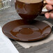 A hand holding a brown Tuxton cappuccino cup over a brown saucer.