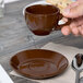 A hand holding a brown Tuxton cappuccino cup over a brown saucer.