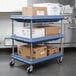 A slate blue Metro utility cart with three deep ledge shelves holding carton boxes.