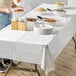 A woman standing at a table with white Hoffmaster tablecloth and plates of food.