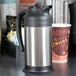 A Choice stainless steel coffee carafe on a counter next to a cup of coffee.