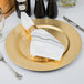 A Tabletop Classics by Walco gold plastic charger plate with silverware and a napkin on a white surface.