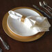 A white Tabletop Classics charger plate with a gold rim on a table with silverware and a white napkin.