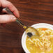 A hand holding a WNA Comet Reflections stainless steel look plastic spoon over a bowl of soup.