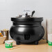 A black Vollrath Colonial Soup Warmer with a lid on a table with cups.