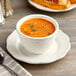 An Acopa ivory stoneware bouillon cup filled with soup on a table with bread and a spoon.