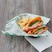 A sandwich with ham and vegetables and chips wrapped in green check deli wrap on a plate.