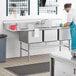 A man in a white shirt and blue apron washing dishes in a Regency stainless steel three compartment sink.