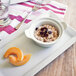 A Tuxton eggshell china casserole dish filled with oatmeal with raisins and a slice of melon on a white plate.