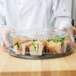 A person in a chef's uniform holding a WNA Comet black round catering tray with sandwiches.