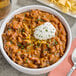 A bowl of Vanee chili with beans with a spoon and a plate of chips.
