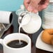 A person pouring cream from a clear plastic creamer into a cup of coffee.