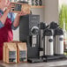 A woman in a red apron using a Bunn black and silver commercial coffee grinder to pour coffee into a bag.
