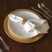 A Tabletop Classics gold plastic charger plate with a white napkin on it.