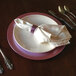 A Tabletop Classics by Walco pink plastic charger plate with a purple rim on a table with silverware and a white napkin.