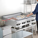 A large stainless steel natural gas countertop griddle with thermostatic controls, shown cooking bacon and pancakes in a commercial kitchen setting.