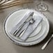 A white Charge It by Jay round charger plate with a silver band on the rim with silverware and a napkin on it.