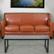 A brown leather Flash Furniture Hercules Diplomat sofa with wooden feet and three pillows in front of a window in a lounge area.