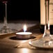 A Leola tea light candle on a table with wine glasses and a wine bottle.