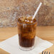 A Cambro clear plastic tumbler with brown liquid, ice, and a straw on a table.