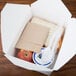 A white Fold-Pak Bio-Pak take-out box on a table with a sandwich, apple, and plastic utensils.