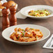 An Acopa ivory stoneware pasta bowl with a plate of pasta and sauce on a table.