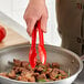 A person using red Mercer Culinary tongs to cook over food.