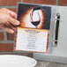 A hand holding a clear menu in front of a white plate on a table.