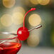 A bright red maraschino cherry with a stem, skewered on a metal pick and garnishing the rim of a cocktail glass.