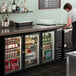 A woman standing at a black Beverage-Air back bar refrigerator full of drinks.