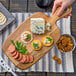 A hand holding a Tablecraft acacia wood display bread board with sliced bread, cheese, and meat on it.