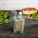 A Tablecraft glass salt shaker with a metal top on a table with a small pile of salt.