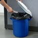A person putting a plastic bag into a blue Rubbermaid BRUTE recycling can with a white lid.