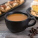 A close-up of a Libbey Driftstone Onyx Satin matte black porcelain cup filled with coffee on a table with pastries.