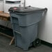 A grey Rubbermaid trash can with wheels and a locking lid next to a printer.