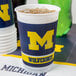 A white Creative Converting plastic cup with a blue and yellow University of Michigan logo and ice in it.