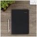 A black At-A-Glance daily appointment book on a wood surface with a pen and a plant.