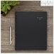 A black At-A-Glance appointment book on a wooden surface with a pen and a plant.