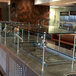 An Advance Tabco stainless steel self service food shield with glass shelves on a large glass counter.
