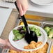 A person holding a black Carlisle polycarbonate solid serving spoon over a plate of food in a salad bar.