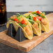 A group of HS Inc. black taco cradles holding tacos with meat and vegetables on a wood board.