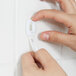 A person's hand using a 3M Command White Mini Hook to hang a white object with a label on a white tile wall.