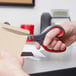 A person using 3M Scotch stainless steel pointed tip scissors with red and dark gray handles to cut a piece of paper.