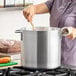 A large aluminum stock pot being stirred with a wooden spoon on a stovetop.