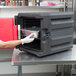 A woman wearing gloves and putting foil containers of food into a black Metro Mightylite food pan carrier with a red door.