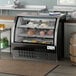 An Avantco black curved glass refrigerated deli case on a counter with meat and cheese inside.