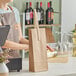 A woman in an apron standing behind a counter with a brown paper wine bag with a bottle in it.