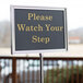 A Chef Master silver hostess/teller sign on a pole with gold text that says "Please Watch Your Step"