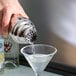 A hand using a Tablecraft stainless steel cobbler shaker to pour a clear liquid into a martini glass.