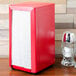 A red Tablecraft Tallfold napkin dispenser on a table with a salt shaker.